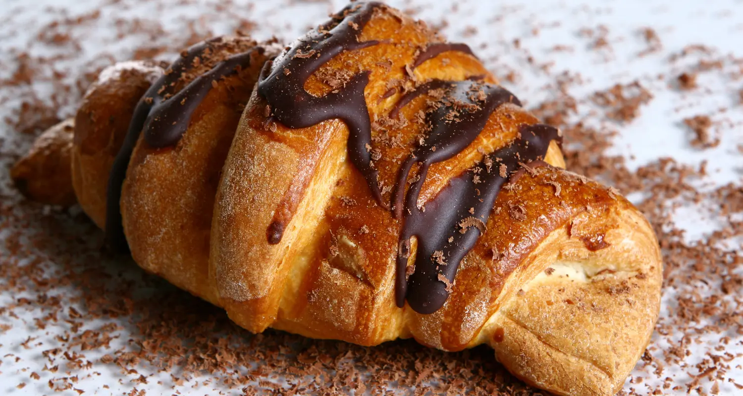 Golden-brown, flaky croissant oozing with Nutella, garnished with a dark chocolate drizzle and scattered chocolate shavings, showcasing a tempting Nutella filled pastry.